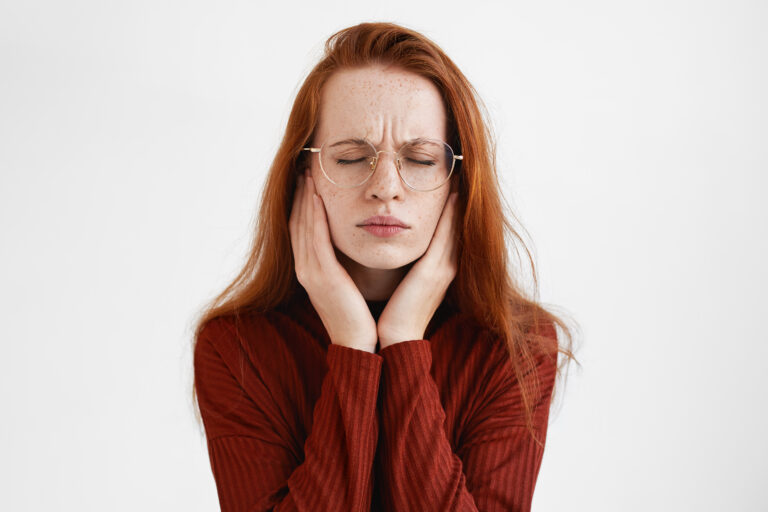 People, health and sickness concept. Portrait of redhead student girl in round spectacles closing eyes and frowning, having painful facial expression, touching face, suffering from toothache